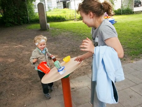 Eine runde Platte auf einem stehenden Rohr dient als Tisch, der gedeckt werden muss. Bei einer Frau und einem kleinen Kind gerät er ins Wackeln und Schüsseln und Flasche fallen runter.