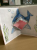 Eine Klappkarte, in der ein gemalter Hai das Maul öffnet.